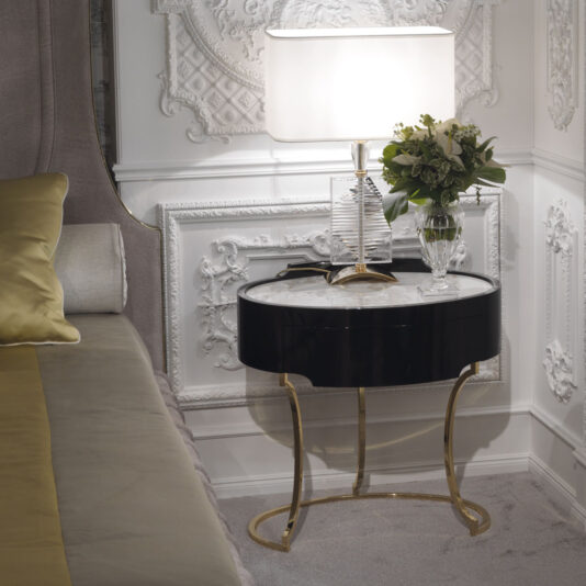 A stylish bedroom features the Oval Italian Ebony Bedside Table with elegant gold legs. On top sits a lamp with a white shade, a clear decorative vase, and a bouquet of flowers. The intricately designed white wall paneling adds a sophisticated touch to the space.