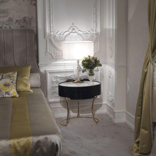 Elegant bedroom featuring a luxurious bed with gray and golden bedding, an Oval Italian Ebony Bedside Table, and a stylish lamp. Ornately decorated wall paneling adds a touch of sophistication, while a vase with fresh flowers enhances the serene ambiance.