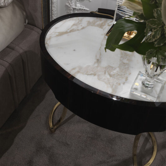 A close-up of a luxurious living room corner features an Oval Italian Ebony Bedside Table with a marble top and gold legs. The table holds a glass vase filled with green leaves and white flowers, positioned next to a plush gray couch on a soft gray carpet.