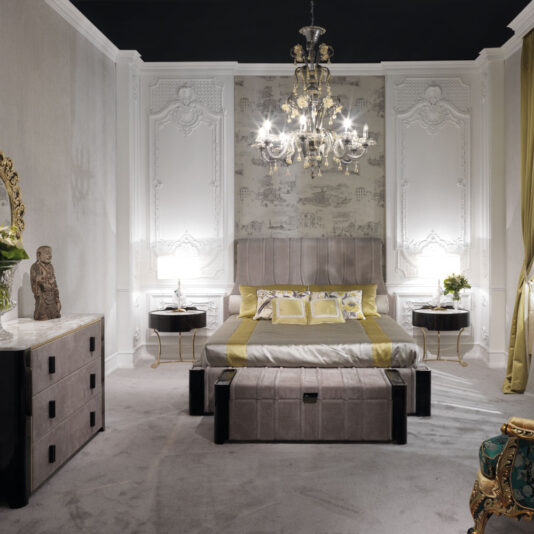 A luxurious bedroom showcasing an elegant chandelier, a spacious bed with a padded headboard, and stylish bedding adorned with yellow accents. The room is embellished with ornate wall paneling, a reflective dresser, two Oval Italian Ebony Bedside Tables complete with lamps, and a decorative statue.