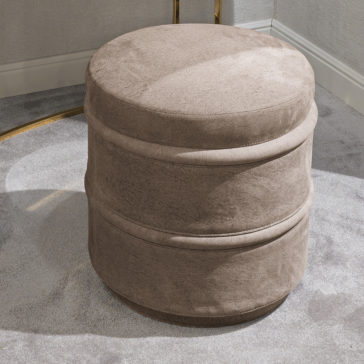 Luxury Leather Footstool The Luxury Leather Footstool, featuring a round shape and taupe-colored luxury leather with a velvet-like texture, rests on a light gray carpet beside a beige wall accented with white trim. The ottoman boasts a minimalist design highlighted by a horizontal seam around its middle.