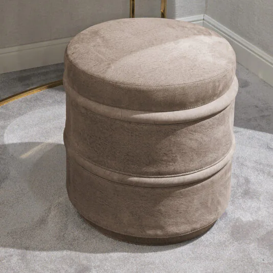 The Luxury Leather Footstool, featuring a round shape and taupe-colored luxury leather with a velvet-like texture, rests on a light gray carpet beside a beige wall accented with white trim. The ottoman boasts a minimalist design highlighted by a horizontal seam around its middle.