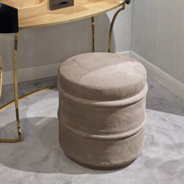 Luxury Leather Footstool A round, beige ottoman with a soft, plush texture sits on a light gray carpet. The ottoman is placed in front of a wooden desk with gold, curved legs, against a neutral-colored wall. The room has modern, minimalist decor accentuated by the presence of the Luxury Leather Footstool.