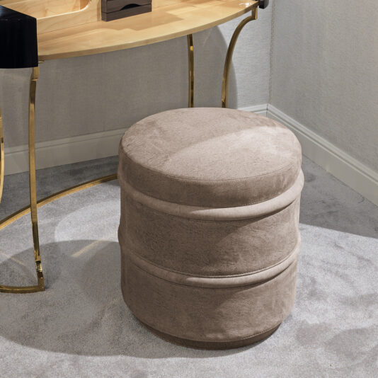 A round, beige ottoman with a soft, plush texture sits on a light gray carpet. The ottoman is placed in front of a wooden desk with gold, curved legs, against a neutral-colored wall. The room has modern, minimalist decor accentuated by the presence of the Luxury Leather Footstool.
