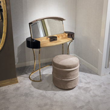 Luxury Leather Footstool A small makeup vanity with a curved mirror is positioned against a wall. The vanity has several compartments for storage and stands on gold legs. In front of it is the Luxury Leather Footstool, featuring light brown, suede-like fabric. The room is carpeted in gray.
