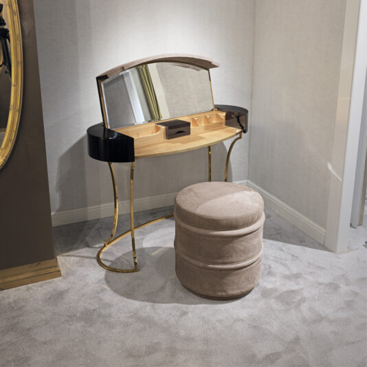 A small makeup vanity with a curved mirror is positioned against a wall. The vanity has several compartments for storage and stands on gold legs. In front of it is the Luxury Leather Footstool, featuring light brown, suede-like fabric. The room is carpeted in gray.