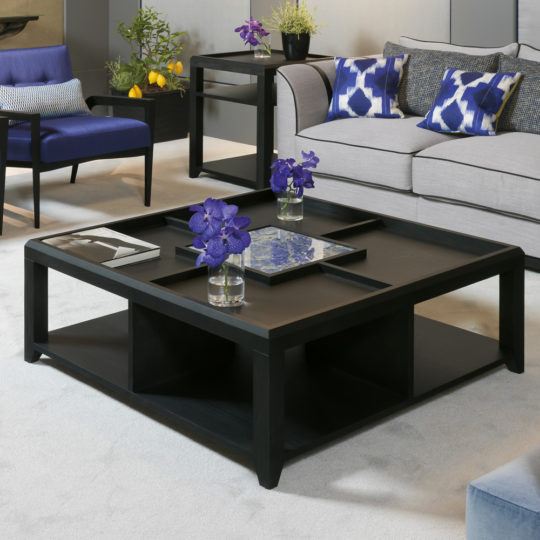 A modern living room is accented by a Luxury Square Coffee Table, featuring geometric compartments, each housing a small glass vase filled with purple flowers. The space also boasts a grey sofa adorned with blue patterned cushions, complemented by a blue armchair. Completing the ensemble is a side table decorated with flowers and a charming plant.
