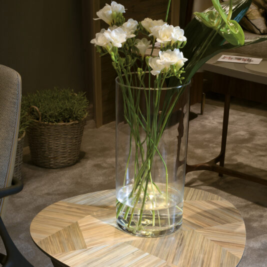 Modern Marquetry Side Table A clear glass vase filled with white flowers and greenery sits on the Modern Marquetry Side Table, which features a round design with a herringbone pattern. The table is placed on a beige carpet, and in the background next to a cushioned chair, there is a wicker basket holding a plant.