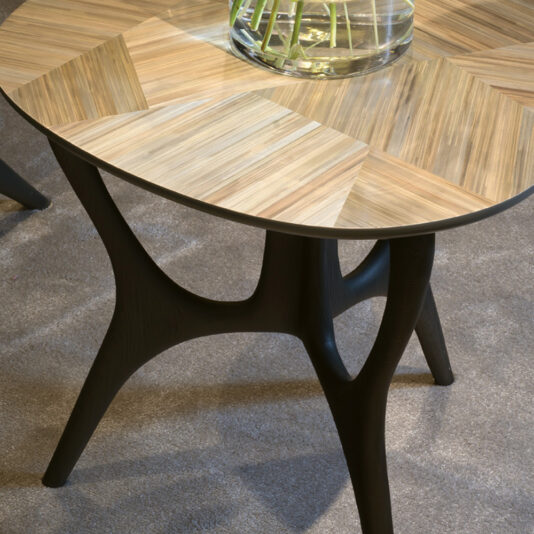 Modern Marquetry Side Table A close-up of the Modern Marquetry Side Table showcases its unique, geometric patterned tabletop. The table features dark wooden legs with sleek, curved lines, all standing on a light-colored carpeted floor. A glass vase with green stems is partially visible on the table.