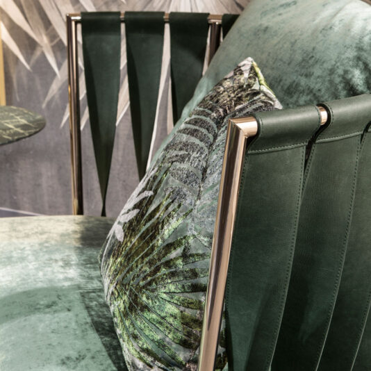 Exclusive Modern Leather Twist Armchair A stylish green velvet chair with a metallic frame and vertical slits on its backrest features a green and white printed cushion adorned with a leafy pattern, evoking the sophisticated vibe of an Exclusive Modern Leather Twist Armchair. The background complements the scene with a wall showcasing a similar leafy design.