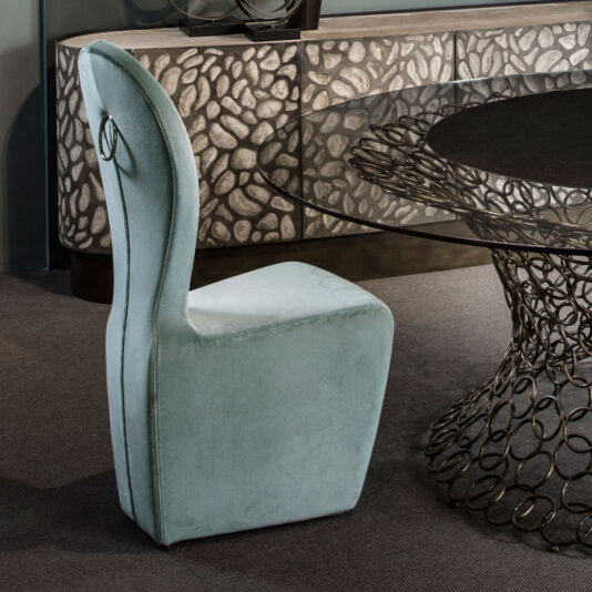 A High Backed Modern Dining Chair, upholstered in plush light blue fabric, stands next to a round glass table with an intricate base made of interlocking metal rings. This arrangement is set in a contemporary room featuring textured stone-like decor on a console in the background.