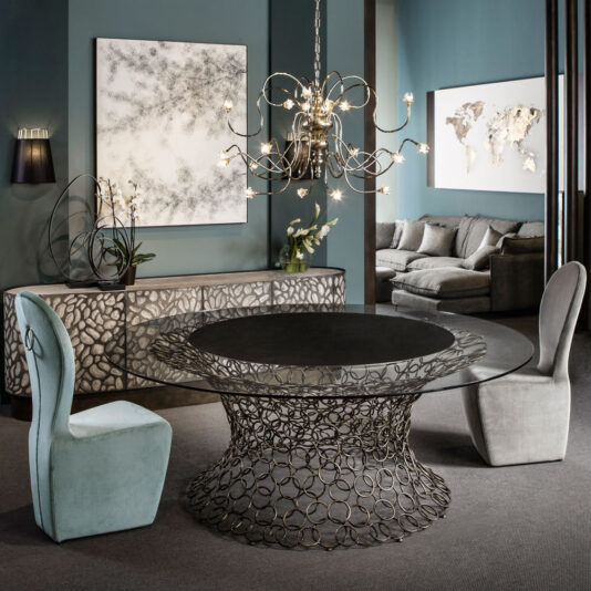 A modern dining room features a round glass table with an intricate metal base, surrounded by high backed modern dining chairs. A stylish chandelier with multiple glowing bulbs hangs above. The walls are adorned with abstract art and a world map, and a sleek sideboard completes the room.
