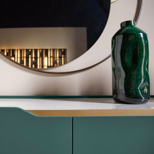 Large Modern Curved Buffet Sideboard A green ceramic vase sits on the Large Modern Curved Buffet Sideboard, which features a green front and a white top. Behind the sideboard, a large round mirror reflects part of the room, offering a glimpse of a decorative element with vertical lines and warm lighting.