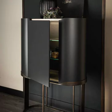 The Large Modern Curved Cabinet features sleek black doors that are partially open, revealing glass shelves inside. It stands on a tall metallic frame against a wall, with decorative items placed on the top shelf and a dark vase with greenery adorning the upper surface.