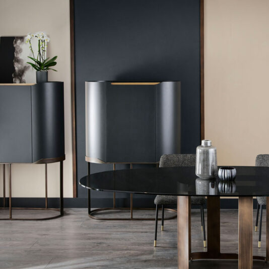 Modern dining room featuring a black-topped circular table with dark wooden legs, surrounded by gray upholstered chairs. In the background, there is a Large Modern Curved Cabinet and another sleek black storage cabinet. A potted orchid and decorative items adorn both cabinets.