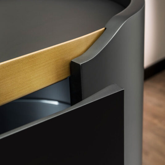 Close-up image of the corner of a Large Modern Curved Cabinet. The design features sleek, black edges with a wooden top. The drawer appears to be partially open, revealing a metallic interior. The overall style is minimalistic and contemporary.