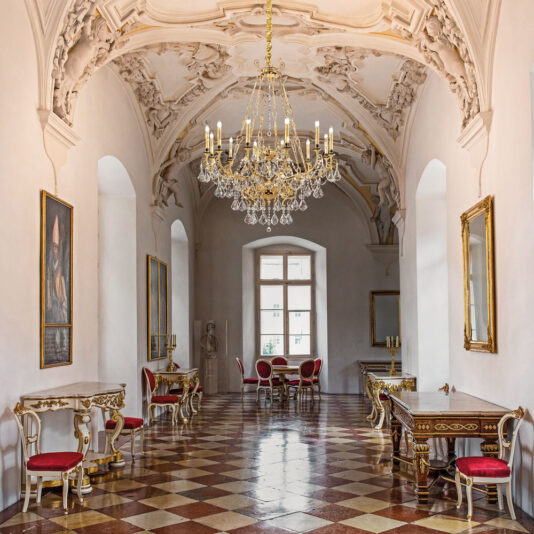 A grand, ornate hall with a high, intricately detailed ceiling featuring a Large Opulent Crystal Pendant Chandelier. The room boasts long windows, decorated walls adorned with framed portraits, elegant red and gold chairs, and wooden tables. The floor is checkered with red and white tiles.