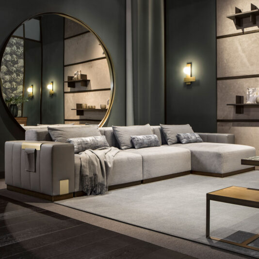A modern living room with a Luxurious Contemporary Modular Corner Sofa in light gray, adorned with cushions and a throw blanket. The space features a circular mirror on the wall, floating shelves with decor, warm wall-mounted lights, and a sleek coffee table on a light gray rug.