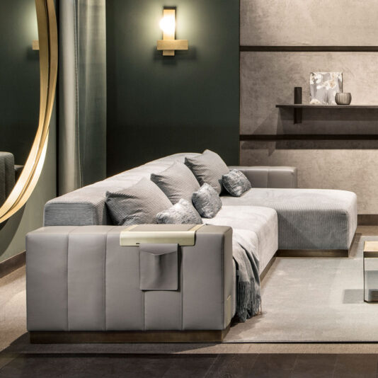 Luxurious Contemporary Modular Corner Sofa A contemporary living room features the Luxurious Contemporary Modular Corner Sofa adorned with gray cushions and a throw, set against a dark green wall. A circular mirror on the wall reflects part of the room. Soft lighting and minimalist decor, including shelves and modern light fixtures, complete the space.