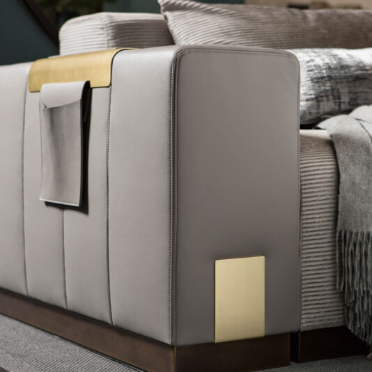 Luxurious Contemporary Modular Corner Sofa A close-up of the Luxurious Contemporary Modular Corner Sofa's armrest, elegantly covered in gray leather and featuring a built-in brass accent. Attached to the armrest is a pocket holding a small item. The sofa is accessorized with a striped cushion and a neatly folded gray blanket, creating an inviting and cozy atmosphere.