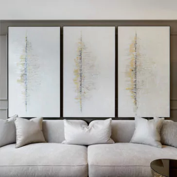 A plush, light gray sofa is positioned in front of a wall adorned with three Luxury Abstract Artwork pieces. Each artwork features a vertical line blended with soft hues of blue, yellow, and white, adding a modern, artistic touch to the room's neutral decor.