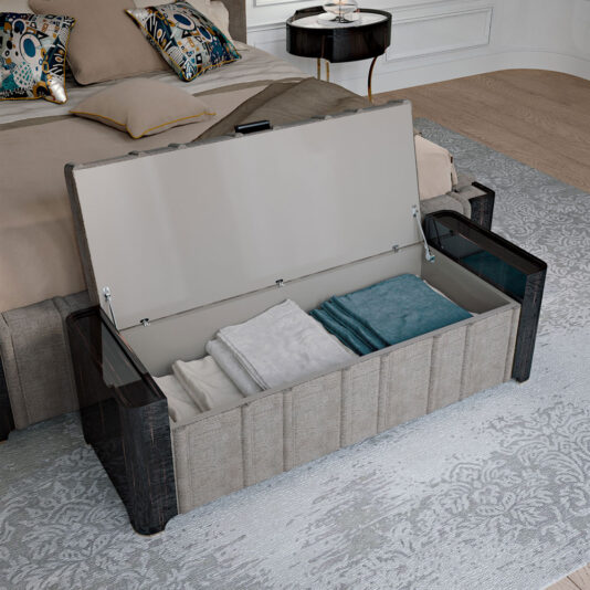 A bedroom featuring the elegant Luxury Ebony Ottoman at the foot of the bed, partially open to reveal neatly folded blankets inside. The bed is adorned with beige bedding and decorative pillows. A side table with a lamp stands beside the bed, and a patterned rug covers the floor.