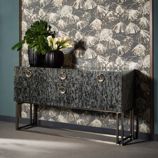 A Luxury Modern Sideboard in dark tones with gold handles stands against a decorative wallpaper featuring a botanical pattern. On top of the sideboard are two black vases containing green leaves and white calla lilies, adding a touch of elegance to the setup.