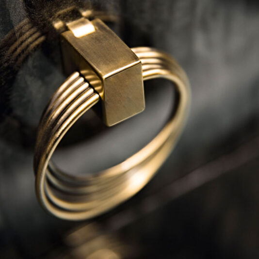 A close-up of a brass door knocker shaped like a ring mounted on a dark-colored door. The ring features a series of concentric circles and is attached to a rectangular bracket, giving the knocker an appearance as modern and elegant as the Luxury Modern Sideboard.