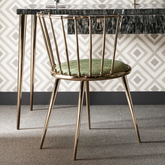 A Luxury Occasional Chair with a gold metal frame and plush green cushioned seat is ideally positioned at a black marble-topped desk, which stands on slender gold legs. The backdrop showcases a geometric white and gray wallpaper pattern, complemented by a textured gray carpet on the floor.