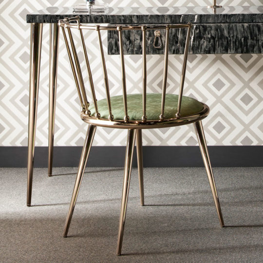 A Luxury Occasional Chair with a gold metal frame and plush green cushioned seat is ideally positioned at a black marble-topped desk, which stands on slender gold legs. The backdrop showcases a geometric white and gray wallpaper pattern, complemented by a textured gray carpet on the floor.
