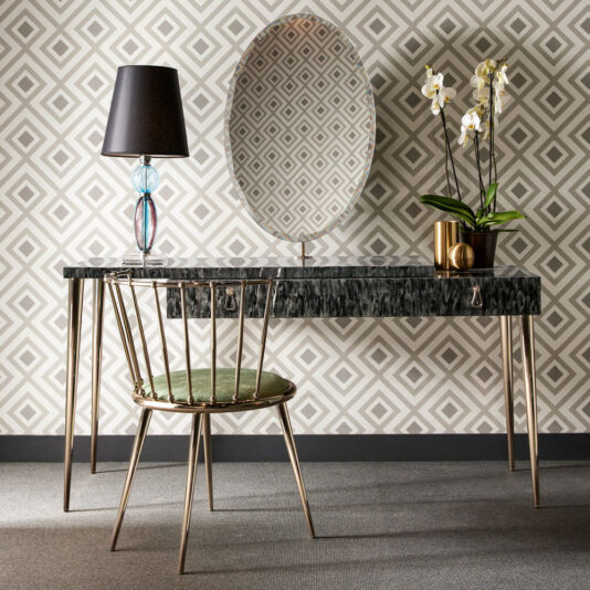 Luxury Occasional Chair A stylish workspace features a black marble desk with gold legs, a Luxury Occasional Chair featuring green cushions, a lamp with a colorful glass base, and a potted orchid. An oval mirror hangs above the desk, all set against geometric patterned wallpaper.