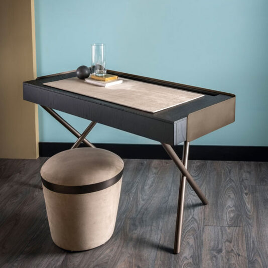 Luxury Round Pouffe A modern desk with a sleek black surface and a beige inset, adorned with items like a glass of water and a book, stands against a light blue wall. Alongside the desk is the Luxury Round Pouffe featuring black accents, all set on a dark wood floor.