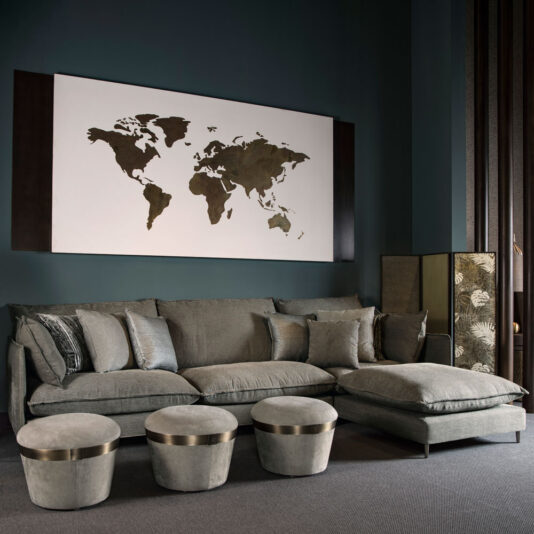 Luxury Round Pouffe In a modern living room set against dark green walls, a plush gray sofa adorned with various gray throw pillows takes center stage. In front of the sofa are three Luxury Round Pouffes featuring elegant metal trims. On the wall behind the sofa hangs a large black and white world map artwork, completing the sophisticated look.