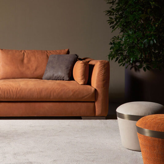 Luxury Round Pouffe A modern living room showcases a brown leather sofa adorned with a dark grey cushion, complemented by a white and orange ottoman and a large potted plant. The inclusion of the Luxury Round Pouffe beautifully enhances the neutral-colored rug and minimal decor, establishing a cozy, sophisticated ambiance.