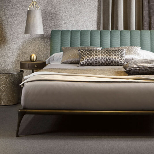 A modern bedroom showcases the Modern Bed With Upholstered Headboard in green, paired with grey and beige pillows and a beige throw blanket. A textured beige side table displays a small gold decorative object, while a hanging lamp with a textured shade illuminates the room. The stylish arrangement is complemented by grey carpeting, emphasizing the contemporary aesthetic of the bed setup.
