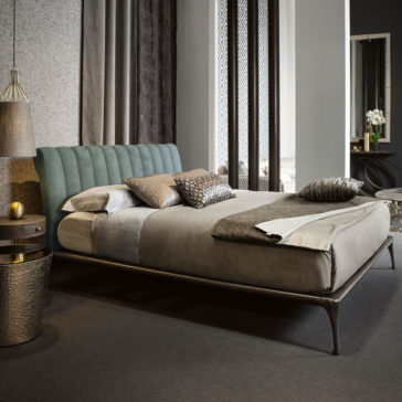 A modern bedroom showcases a cozy Modern Bed With Upholstered Headboard in soft blue, complemented by neutral bedding and decorative pillows. On one side of the bed is a textured round nightstand with a hanging pendant light above. Curtains and a plush carpet complete the serene ambiance.