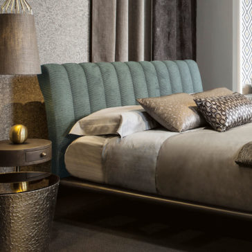 A contemporary bedroom showcasing the Modern Bed With Upholstered Headboard in teal, complemented by grey bedding accented with patterned pillows. A round, metallic bedside table supports a lamp with a textured base and an elegant gold decorative item. The warm lighting enhances the cozy ambiance.