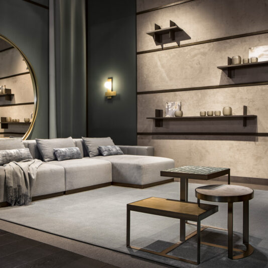 Modern Coffee Table Set A modern living room features a light gray sectional sofa adorned with cushions and a cozy throw. Floating shelves on the wall display decorative items, and a round mirror adds to the aesthetic. The centerpiece is the Modern Coffee Table Set of nested tables positioned in front of the sofa, all contributing to the room's minimalist design characterized by muted colors.