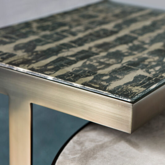 Modern Coffee Table Set A close-up image of the Modern Coffee Table Set corner. The tabletop showcases a textured, abstract design with a black and beige pattern. The table edge is composed of metallic material with a brushed finish, supported by a sturdy, angular leg. The background is blurred.