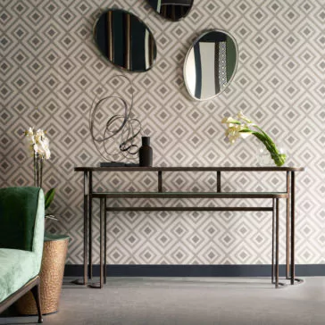 Modern Console Table Set A contemporary living room showcases a geometric pattern wallpaper, a Modern Console Table Set with a sleek metal frame and glass top, and two nested tables tucked beneath it. Three abstract mirrors are mounted above the table, complemented by an orchid in a vase and an abstract sculpture decoratively placed on the tabletop.