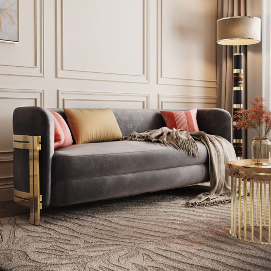 A chic living room showcases the Modern Curved Sofa With Brass Frame, adorned with pink and gold cushions and a beige throw. The room includes a gold-accented floor lamp, a round wooden side table, and a vase with dried flowers. The wall features decorative paneling, and the floor is carpeted.