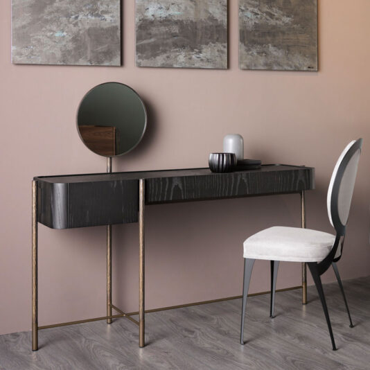 A contemporary vanity arrangement showcasing the **Modern Dressing Table** in dark wood with elegant thin gold legs and a small round mirror, complemented by stylish decorative items. A light beige cushioned chair with a black frame is placed in front of the table. The backdrop features a pinkish-beige wall adorned with abstract art.