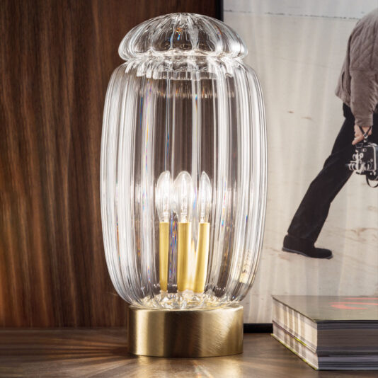 A sophisticated table lamp boasting a ribbed, cylindrical clear glass shade and sturdy brass base, accentuated by its exposed light bulbs. It's elegantly positioned on a wooden surface next to a stack of books and the Modern Curved Sofa With Brass Frame, with a framed photo partially visible in the background.