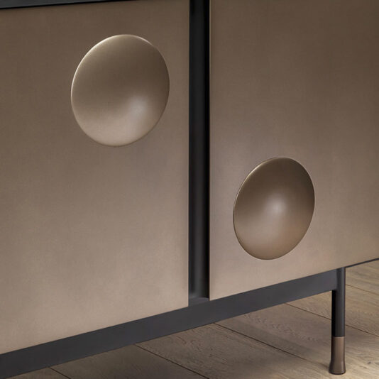 Modern Industrial 4 Door Sideboard A close-up view of the Modern Industrial 4 Door Sideboard showcases its sleek metallic finish. The cabinet doors are adorned with two concave circular indentations, contributing a distinctive design element. The sideboard stands on black legs, with a light wooden floor visible underneath.