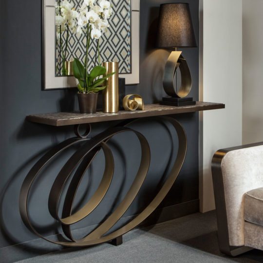 A modern luxury hallway features the Modern Luxury Swirl Console with abstract circular metal legs, adorned with an orchid plant, a gold vase, and an elegant decorative piece. Above the console is patterned wall art, and to the side is a contemporary lamp with a dark shade, beside a stylish beige chair.