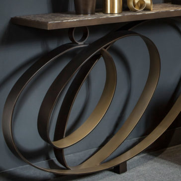 The Modern Luxury Swirl Console, featuring a dark marble top and a distinctive base design with large, overlapping metal rings, is elegantly adorned with metallic vases and sculptures. It is set against a dark wall background and rests on a gray carpeted floor.