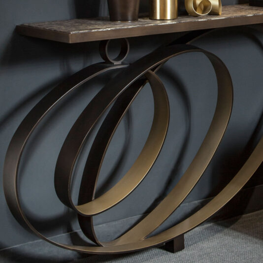 The Modern Luxury Swirl Console, featuring a dark marble top and a distinctive base design with large, overlapping metal rings, is elegantly adorned with metallic vases and sculptures. It is set against a dark wall background and rests on a gray carpeted floor.