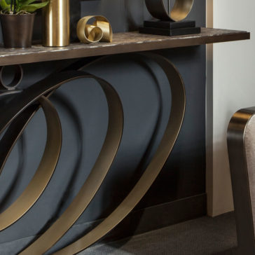 The Modern Luxury Swirl Console features a sleek dark brown surface and distinctive intertwined brass metal legs. It is beautifully adorned with decorative items including a potted plant, a gold sculpture, and a small square black pedestal. The surrounding wall and adjacent furniture maintain a simple and neutral aesthetic.