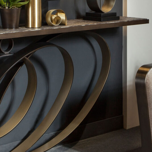 The Modern Luxury Swirl Console features a sleek dark brown surface and distinctive intertwined brass metal legs. It is beautifully adorned with decorative items including a potted plant, a gold sculpture, and a small square black pedestal. The surrounding wall and adjacent furniture maintain a simple and neutral aesthetic.
