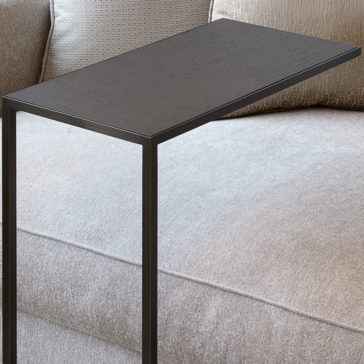 Contemporary Designer Over Sofa Side Table The Contemporary Designer Over Sofa Side Table, featuring a sleek black minimalist metal frame, is positioned over the arm of a light grey sofa. This stylish addition hovers above the seating area, offering a convenient surface for placing items. The sofa itself boasts textured fabric and beige cushions.